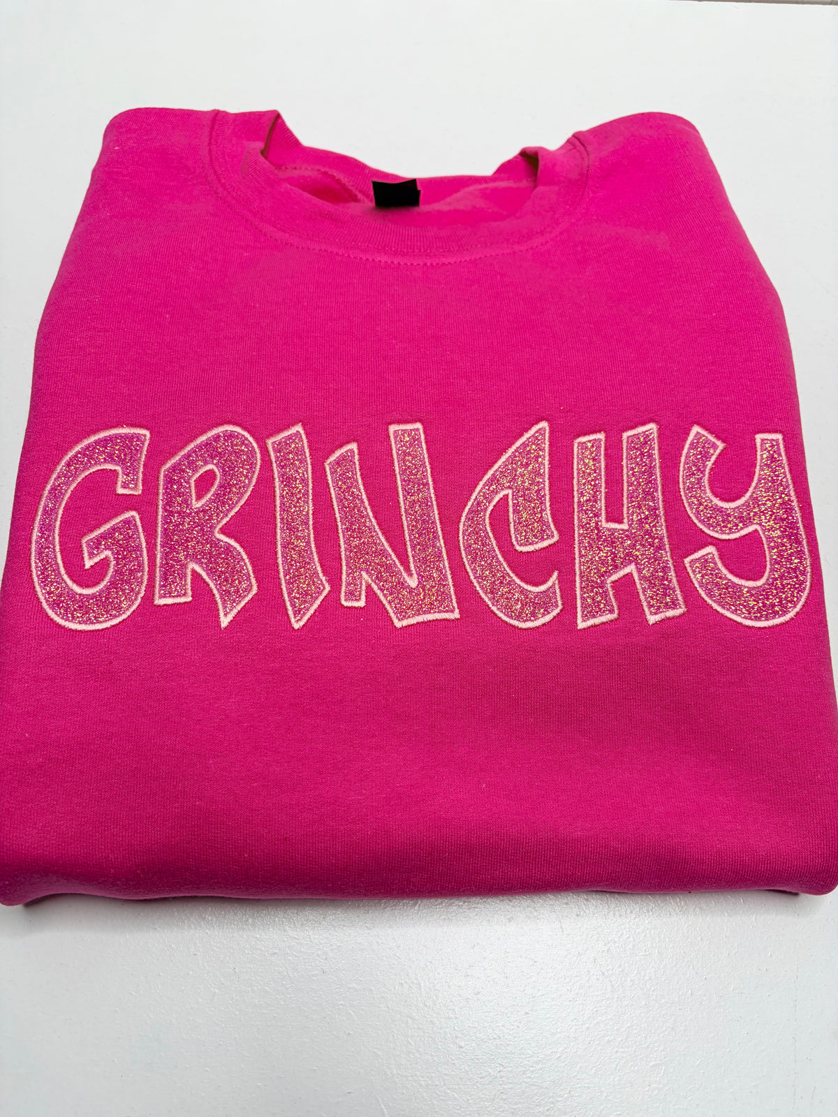 A folded hot pink sweatshirt featuring the word 'GRINCHY' printed across the chest in a distressed, light pink outline font.