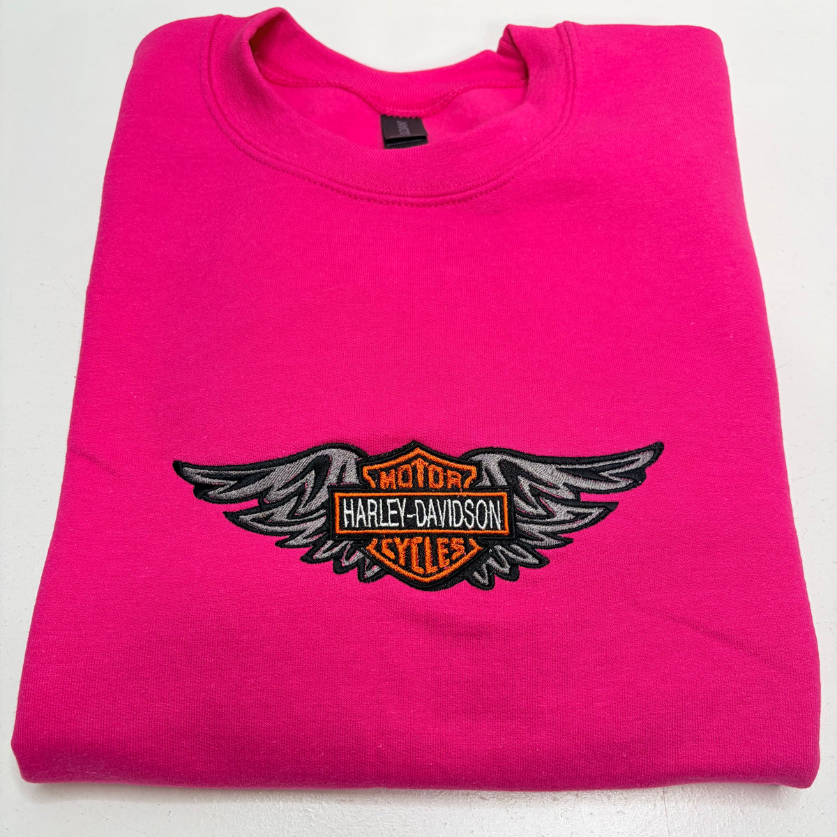 A folded hot pink sweatshirt featuring an embroidered Harley-Davidson winged bar logo on the chest, displayed on a clean white background.