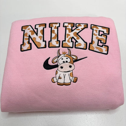 A folded light pink sweatshirt featuring a large arched 'NIKE' graphic in a brown-and-white cow-print pattern, with a cartoon cow and black swooshes underneath, on a plain white background.