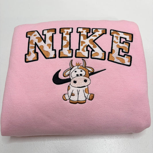 A folded light pink sweatshirt featuring a large arched 'NIKE' graphic in a brown-and-white cow-print pattern, with a cartoon cow and black swooshes underneath, on a plain white background.