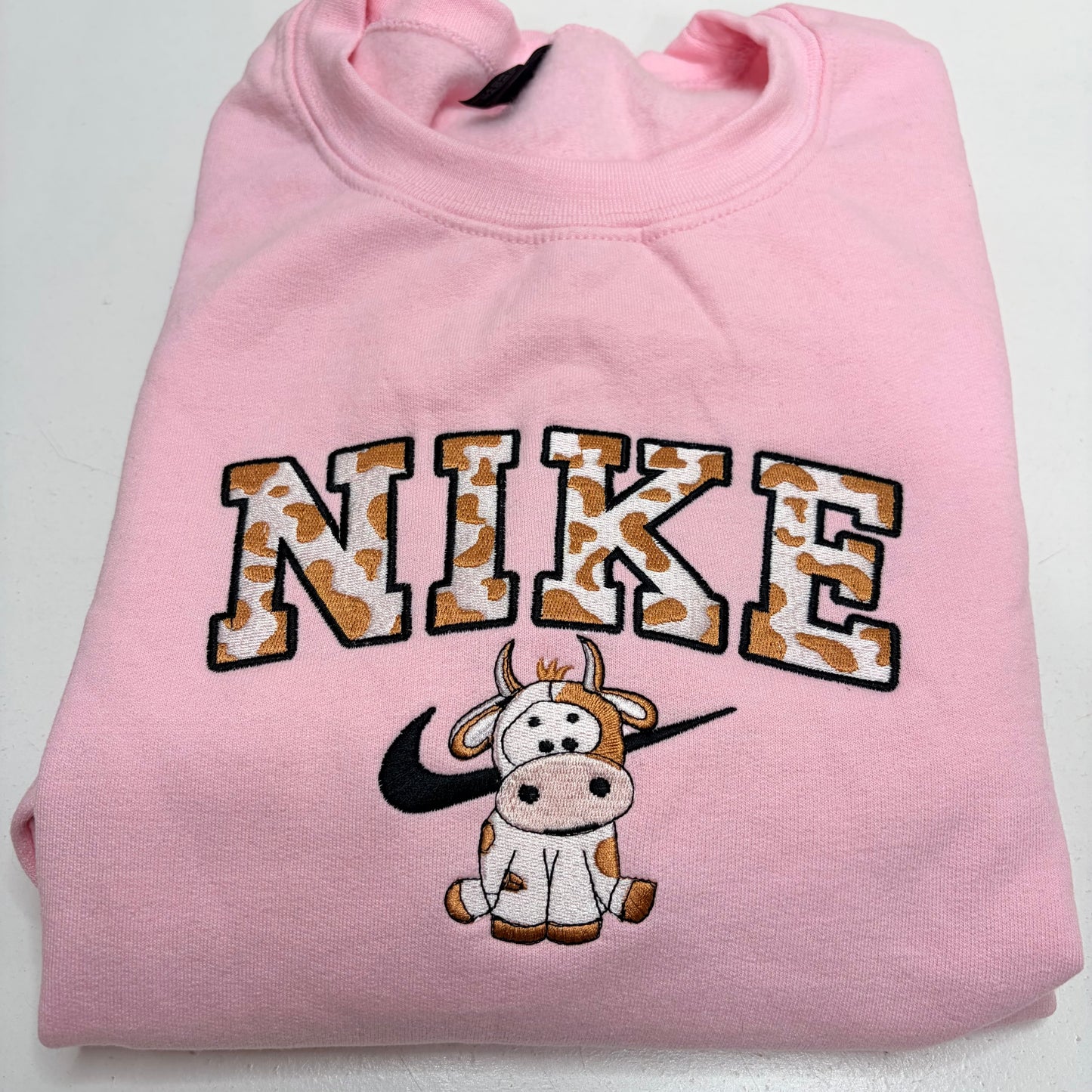 A folded light pink sweatshirt featuring a large embroidered 'NIKE' logo in a cow-print pattern with black outlines, a black Nike swoosh, and a cartoon cow graphic below.
