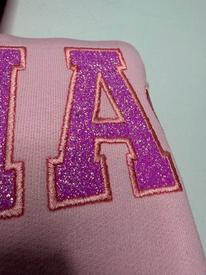 A close-up of a pink fabric item featuring large glittery purple letters outlined in pink thread.
