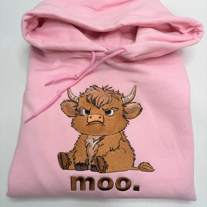 A light pink hoodie featuring a cartoon illustration of an angry brown cow with the word 'moo.' embroidered below it, shown folded on a white background.