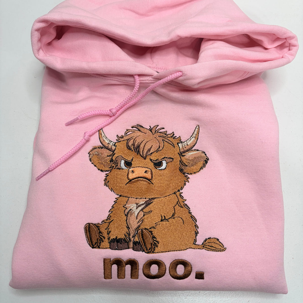light pink hooded sweatshirt with an embroidered grumpy looking scottish highland cow with the word moo under it