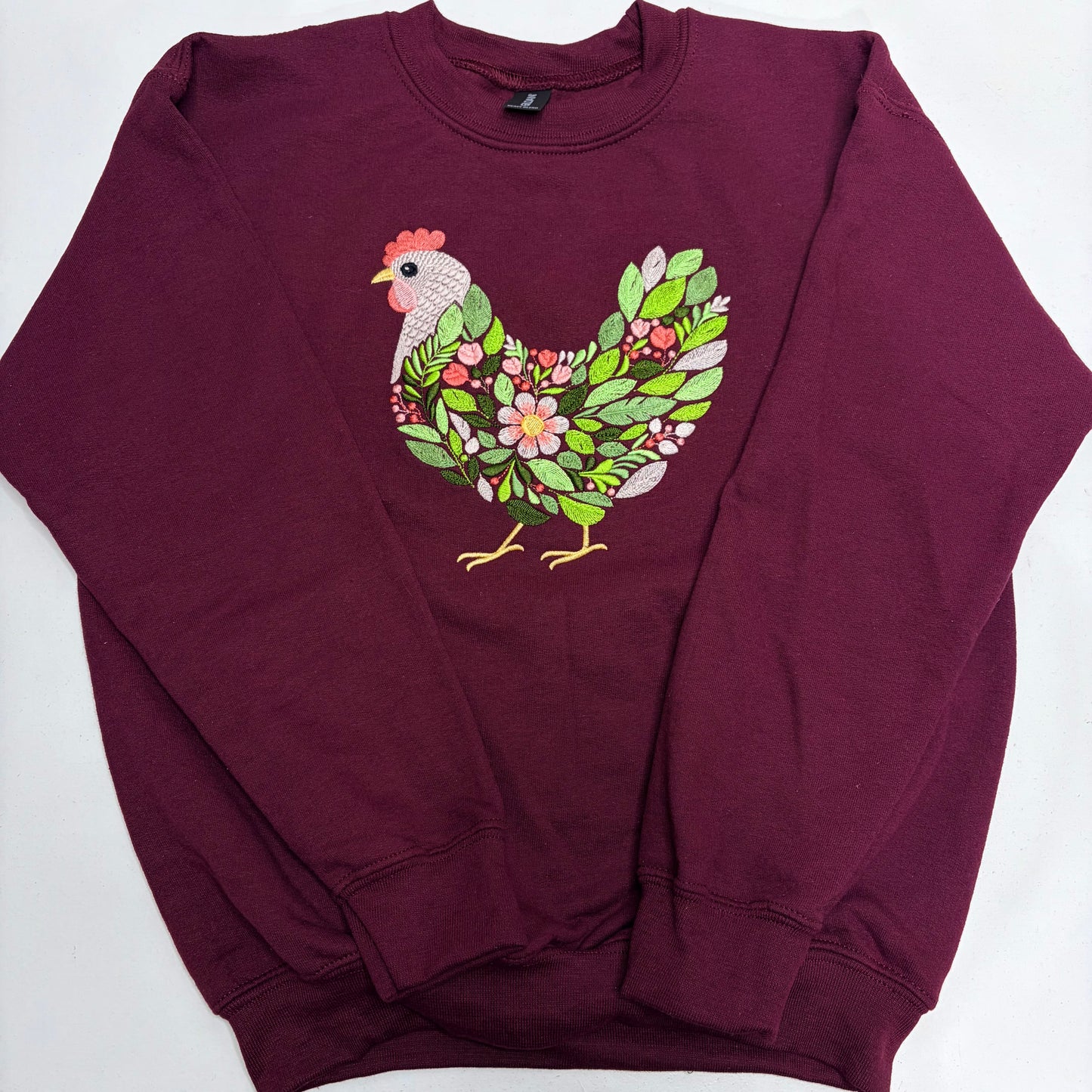 A maroon crewneck sweatshirt featuring a graphic of a chicken filled with green leaves and pink flowers on the chest, laid flat on a white background.