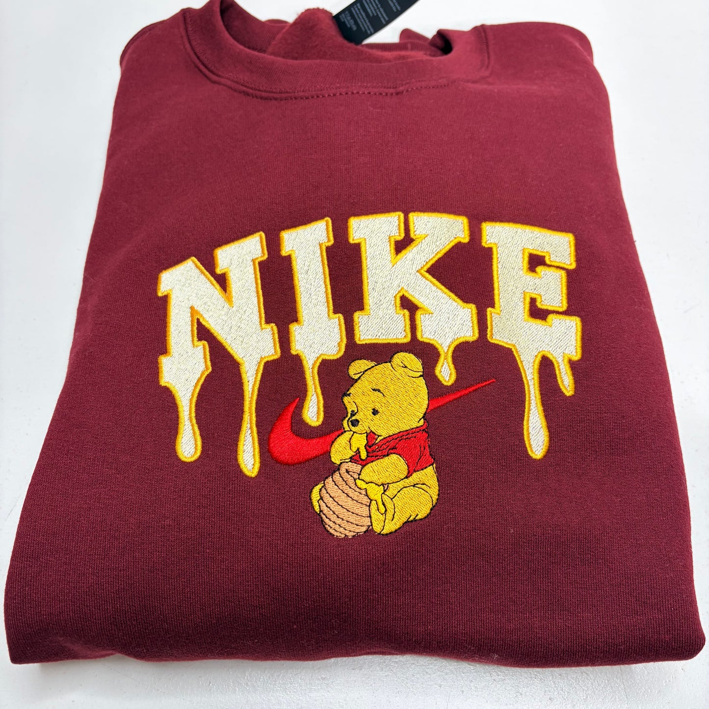 A folded maroon sweatshirt featuring a large cream and yellow dripping 'NIKE' graphic with a red swoosh and winnie the pooh bear holding a honey pot underneath.