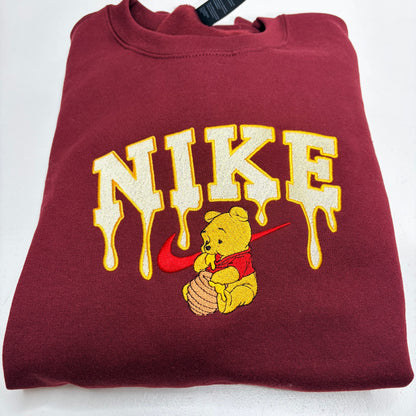 A folded maroon sweatshirt featuring a large cream and yellow dripping 'NIKE' graphic with a red swoosh and winnie the pooh bear holding a honey pot underneath.