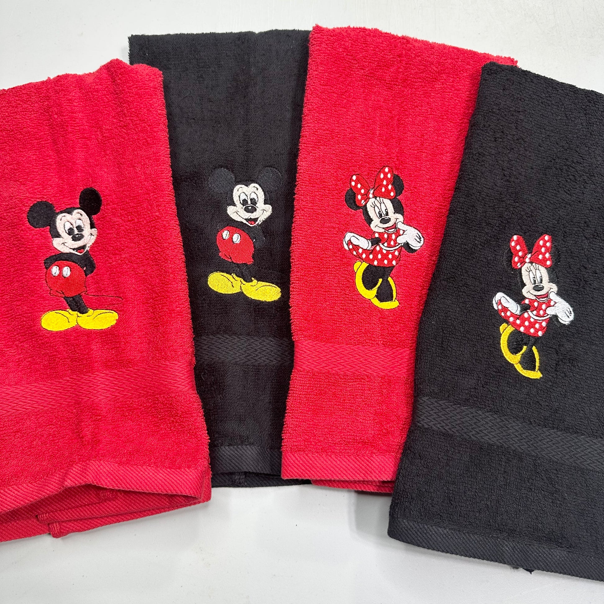 Four folded towels arranged in a fan shape on a light gray background, alternating between red and black, each embroidered with a Mickey Mouse or Minnie Mouse character design.