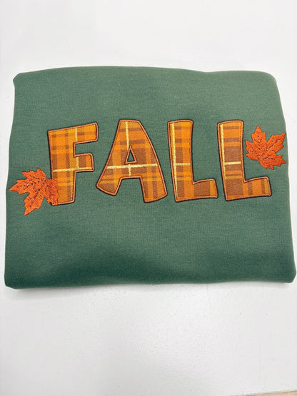 A folded green sweatshirt featuring the word 'FALL' in orange and brown plaid letters, accented with two orange maple leaves on either side, displayed on a white background.
