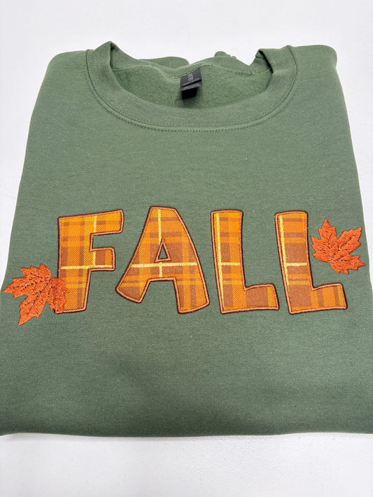An olive green crewneck sweatshirt folded flat, featuring the word 'FALL' in orange plaid letters with orange maple leaf accents on either side, displayed on a white background.