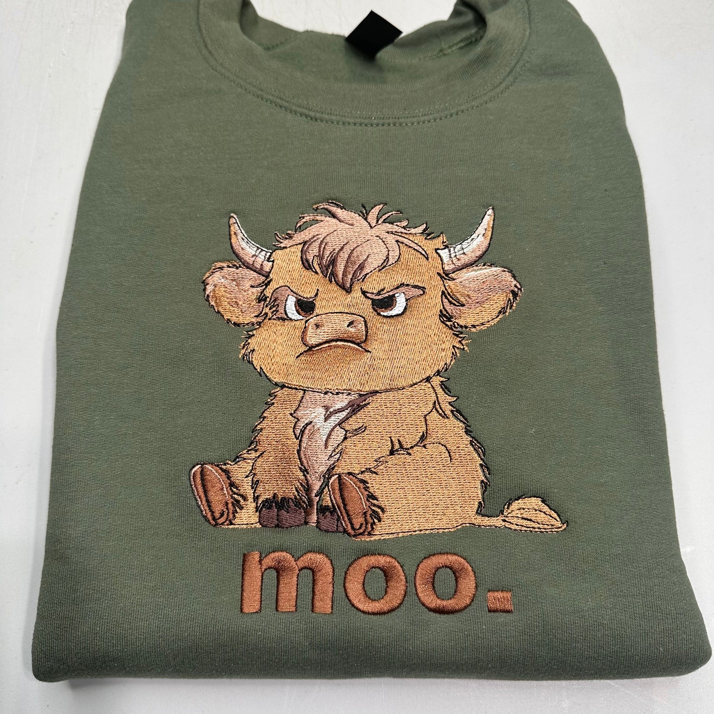 An olive green folded sweatshirt featuring a detailed embroidered cartoon highland cow with the word 'moo.' stitched below it, set against a plain white background.