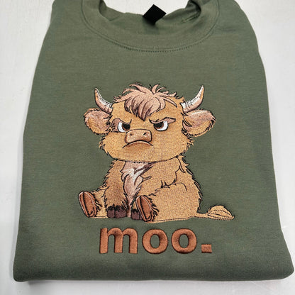 An olive green folded sweatshirt featuring a detailed embroidered cartoon highland cow with the word 'moo.' stitched below it, set against a plain white background.