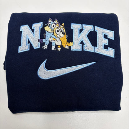 A folded navy blue sweatshirt featuring a large embroidered 'NIKE' logo with two cartoon bluey characters inside the letters and a silver swoosh below, shown on a plain white background.