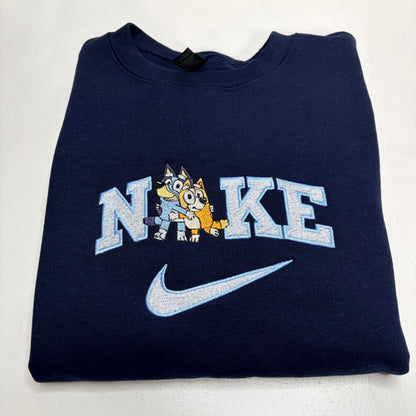 A folded navy blue Nike sweatshirt featuring a light blue and white 'NIKE' graphic with two cartoon bluey bingo dogs in the center and a light blue swoosh below, shown on a plain white background.