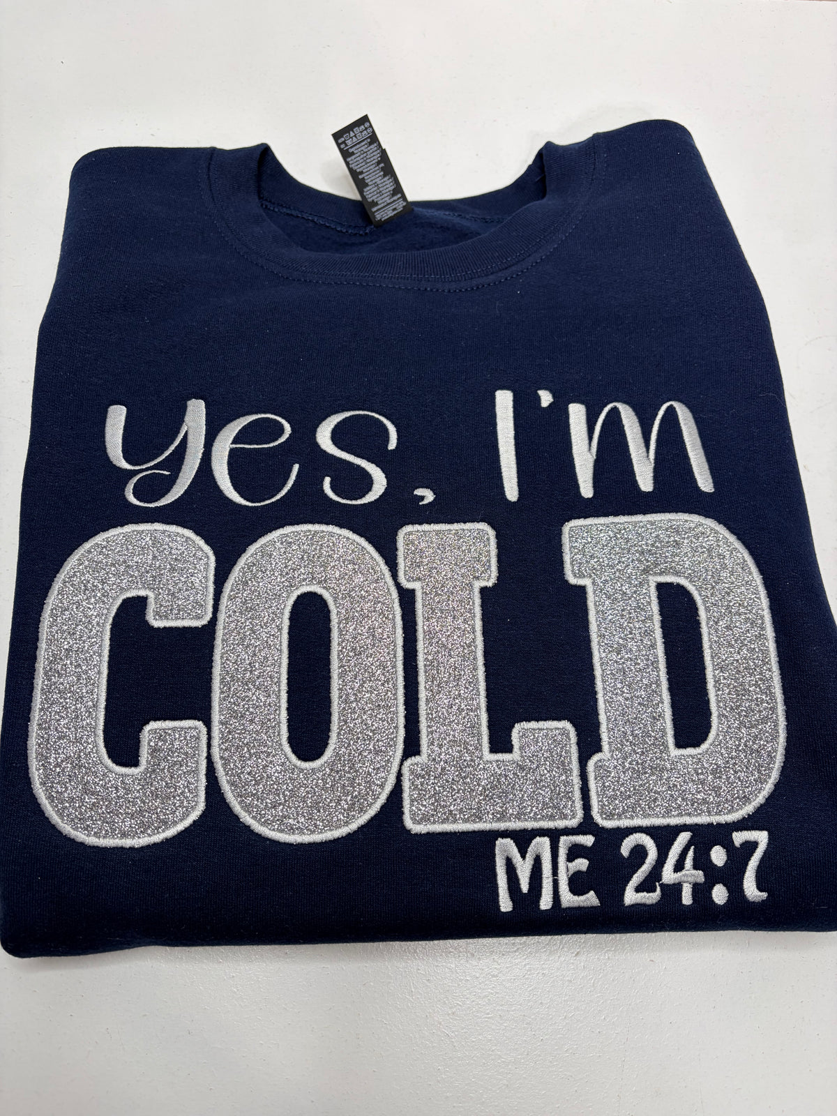 A folded navy blue sweatshirt with 'Yes, I'm COLD ME 24:7' embroidered in white and silver glitter lettering on the front, displayed on a light gray background.