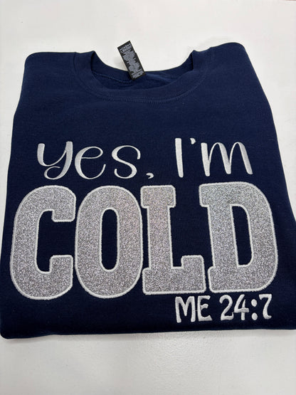 A folded navy blue sweatshirt with 'Yes, I'm COLD ME 24:7' embroidered in white and silver glitter lettering on the front, displayed on a light gray background.