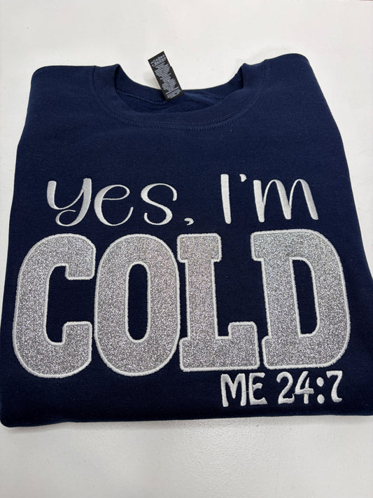 A folded navy blue sweatshirt with 'Yes, I'm COLD ME 24:7' embroidered in white and silver glitter lettering on the front, displayed on a light gray background.