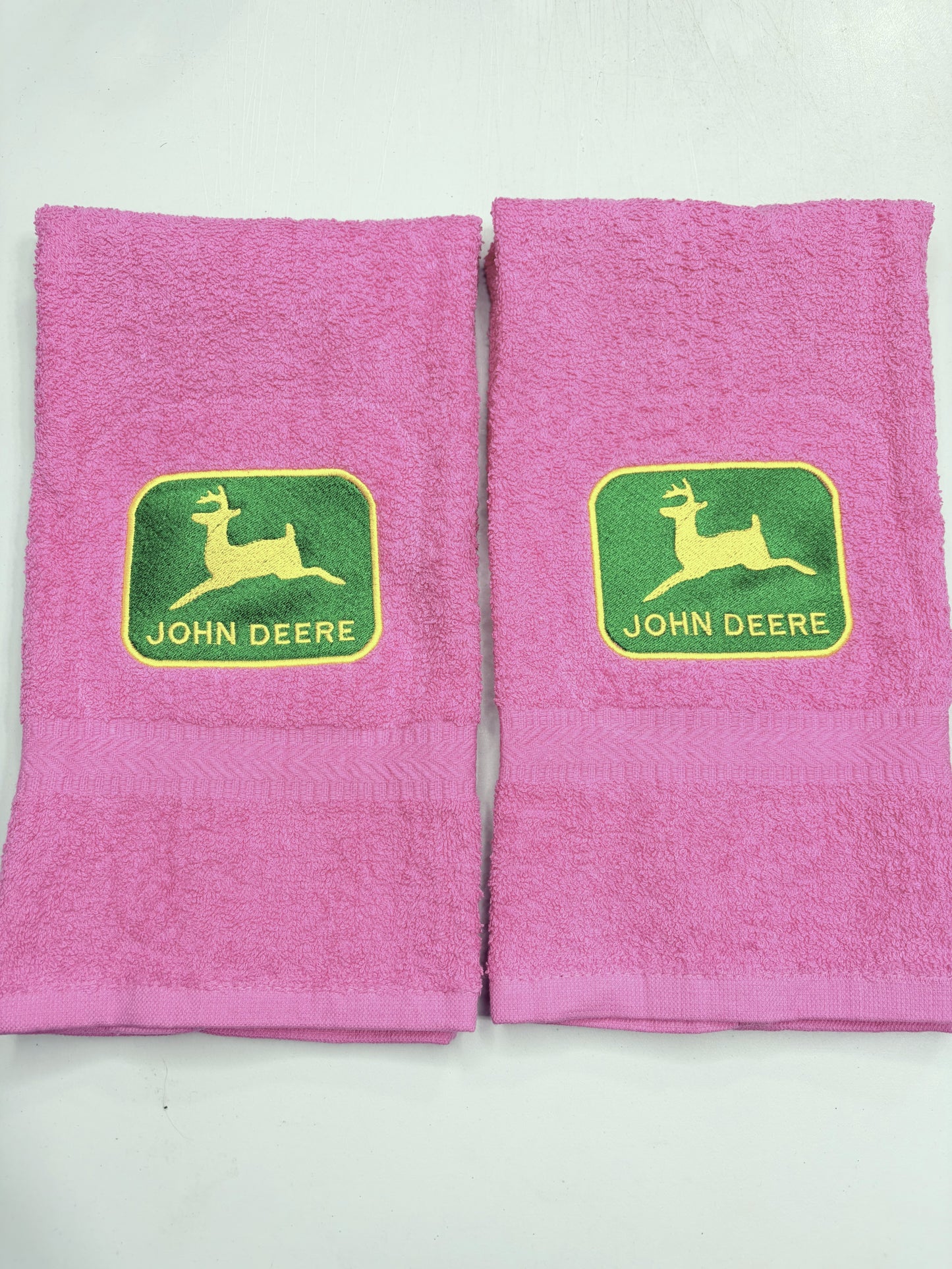 Two pink towels with green and yellow John Deere logos on the front, displayed side-by-side on a white background.