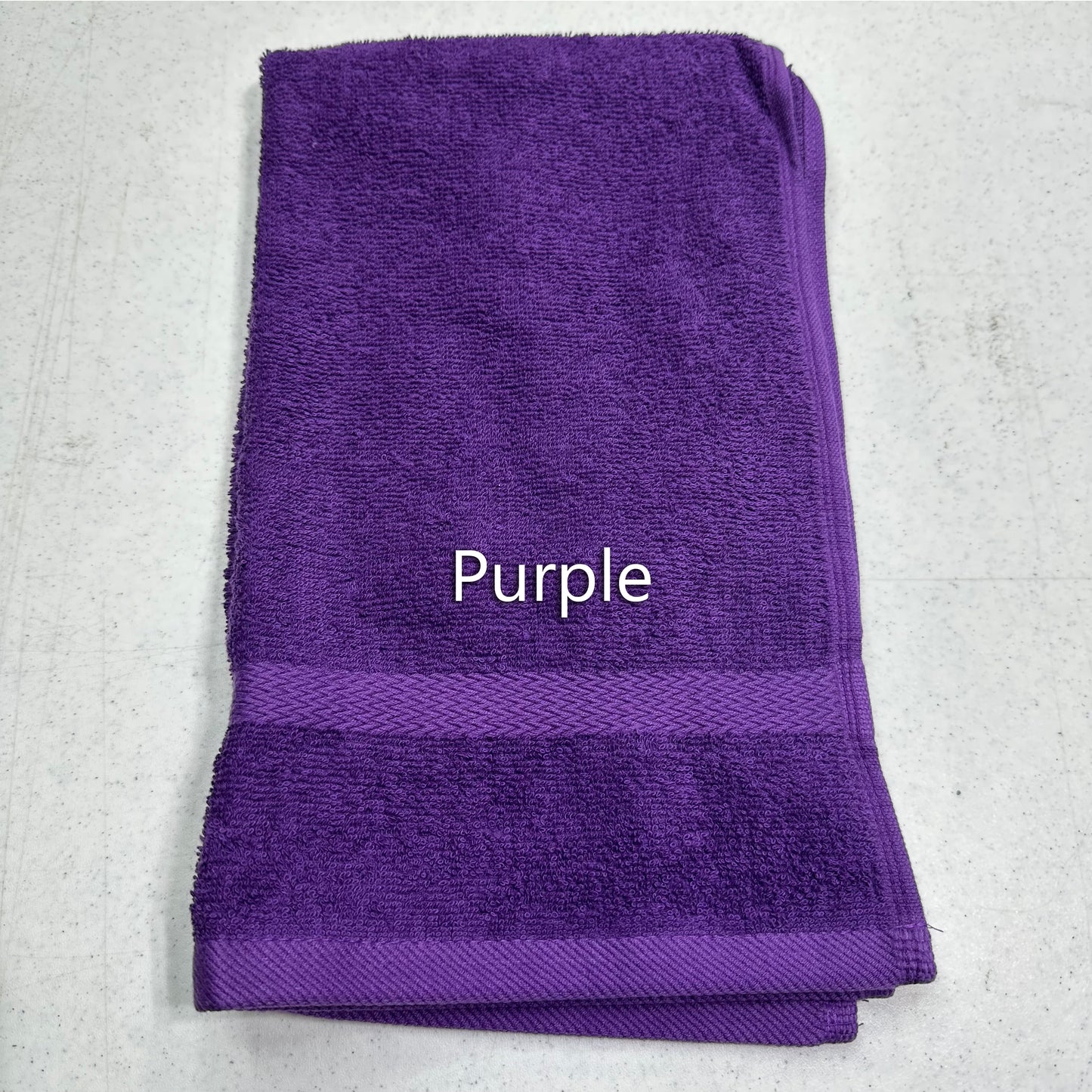 A folded purple terry cloth towel with a woven border and the word 'Purple' printed in white text in the center, laid flat on a light gray textured surface.