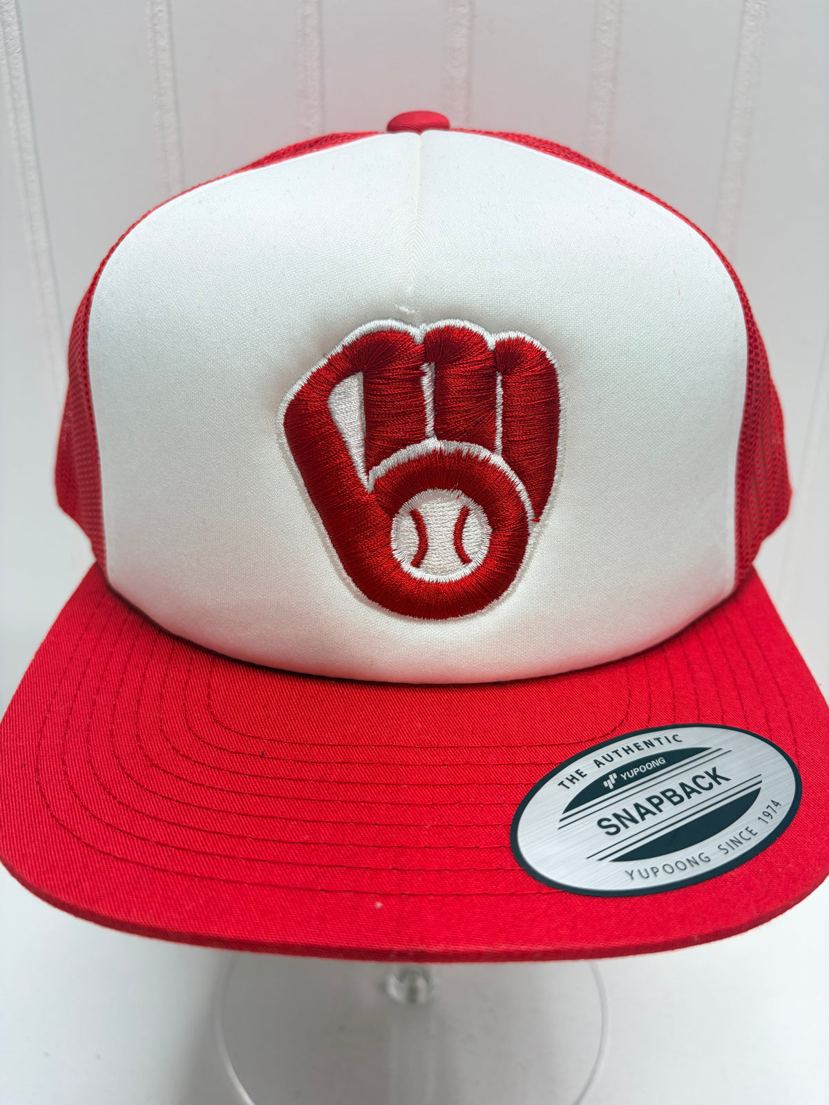 A red and white snapback trucker hat featuring an embroidered baseball glove and baseball logo on the front panel, displayed on a clear stand against a white paneled background.