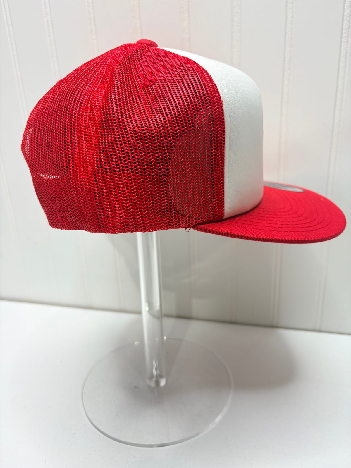 A red and white trucker cap with a red mesh back and red flat brim, displayed on a clear acrylic stand against a white paneled background.