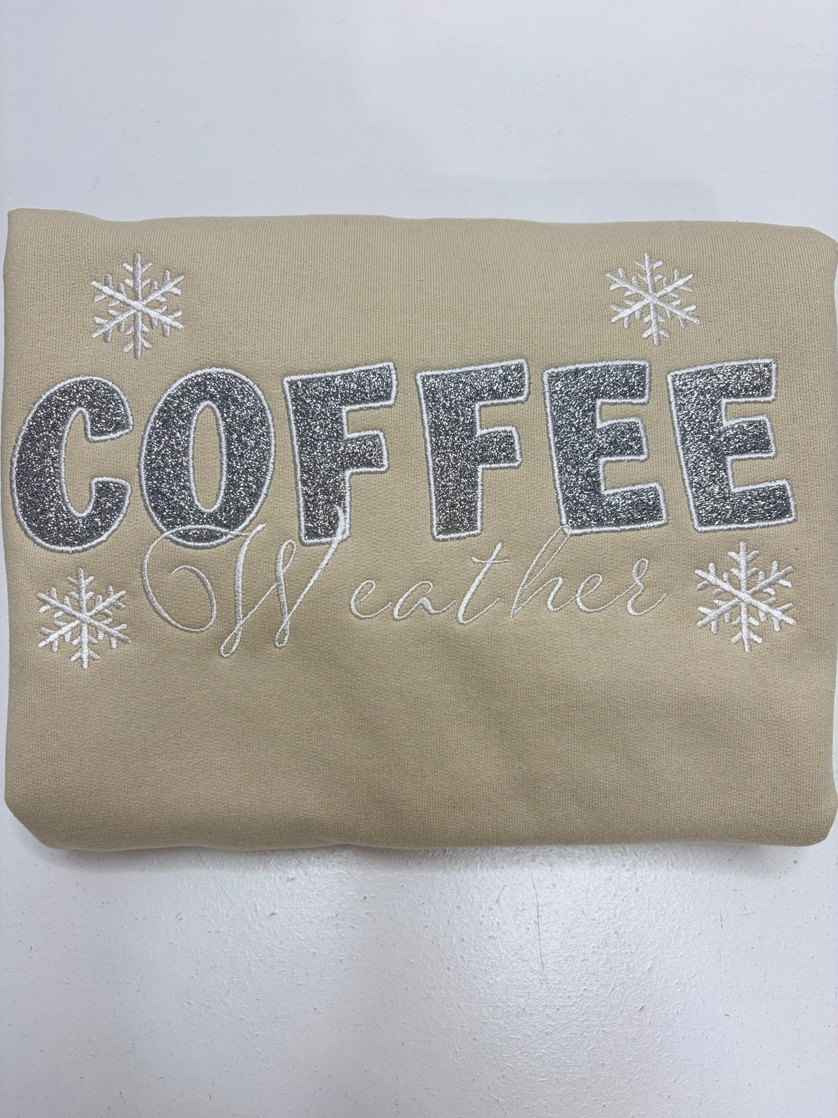 A beige sweatshirt with 'COFFEE weather' printed in silver glitter and white script, decorated with white snowflakes, shown folded on a white background.