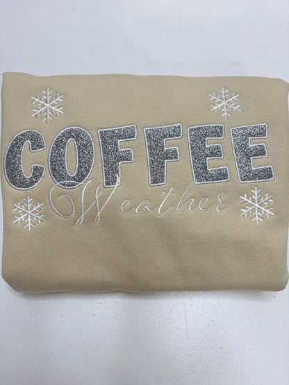 A beige sweatshirt with 'COFFEE weather' printed in silver glitter and white script, decorated with white snowflakes, shown folded on a white background.