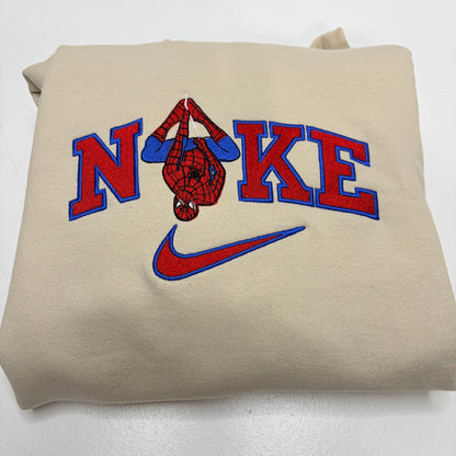 A folded beige sweatshirt featuring a red and blue embroidered design that spells 'NOKE' with a Spider-Man graphic inside the letters and a Nike swoosh below.