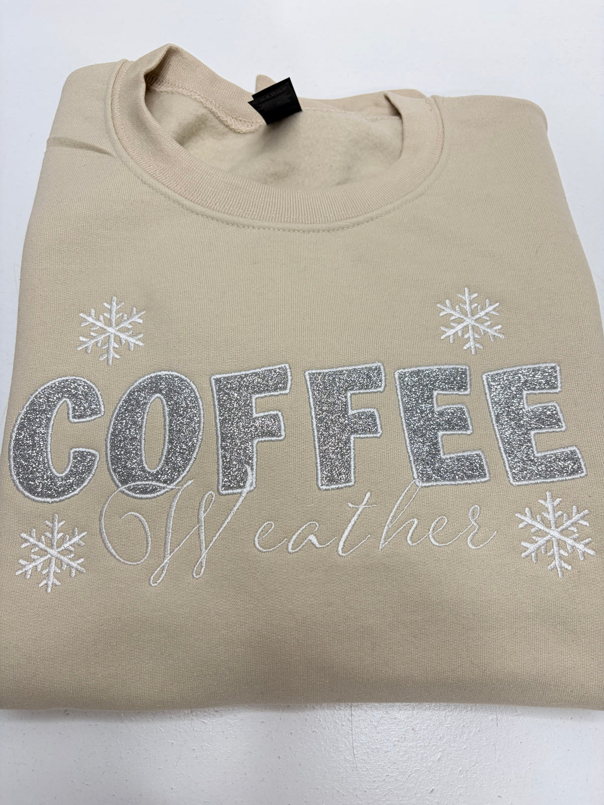 A beige crewneck sweatshirt with 'COFFEE weather' printed in silver glitter and script, decorated with silver snowflakes, laid flat on a white background.