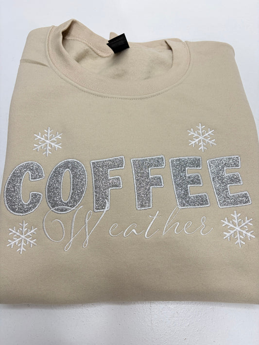 A beige crewneck sweatshirt with 'COFFEE weather' printed in silver glitter and script, decorated with silver snowflakes, laid flat on a white background.