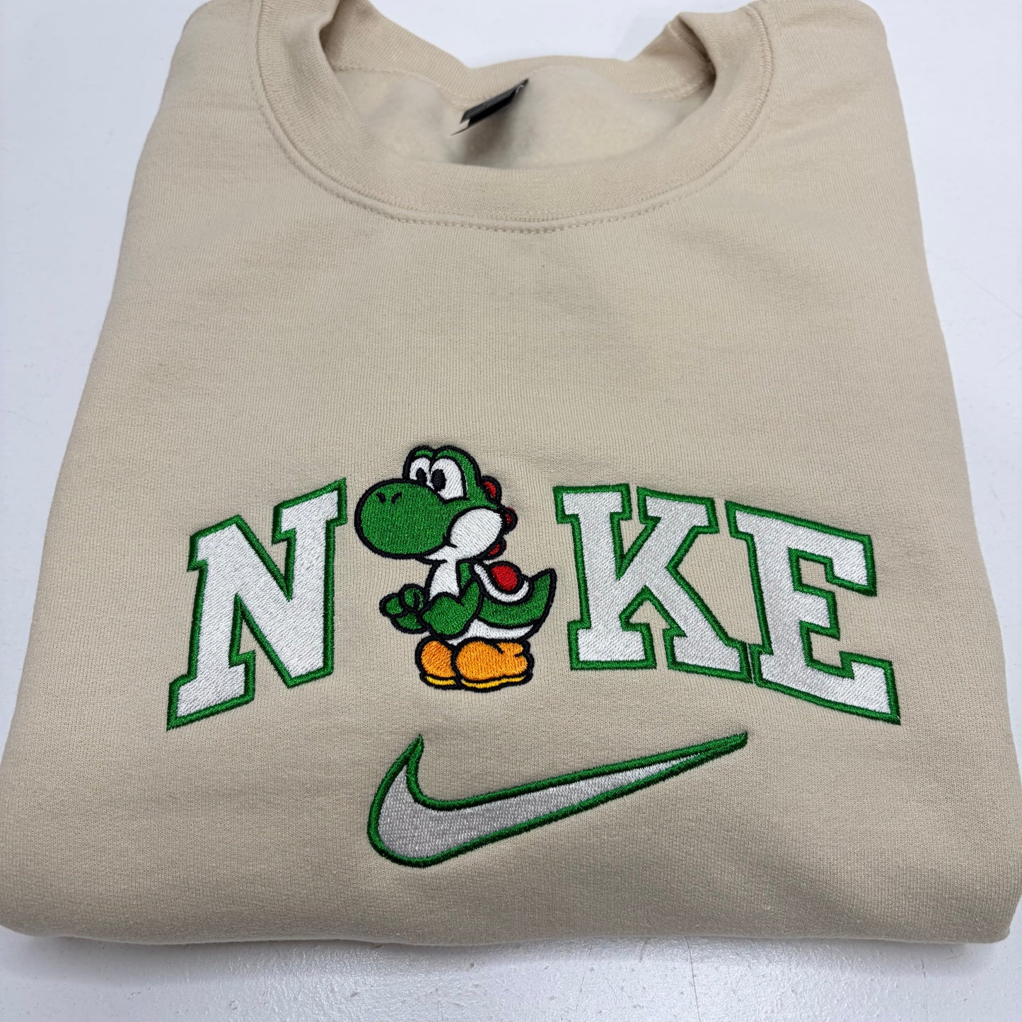 A beige crewneck sweatshirt featuring a custom 'Nike' logo design with a green and white Yoshi character and a green embroidered swoosh on the front.