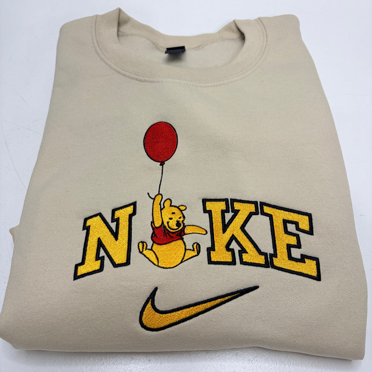 A beige sweatshirt featuring a custom 'Nike' design with a embroidered Winnie the Pooh holding a red balloon, displayed on a white background.