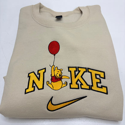 A beige sweatshirt featuring a custom 'Nike' design with a embroidered Winnie the Pooh holding a red balloon, displayed on a white background.