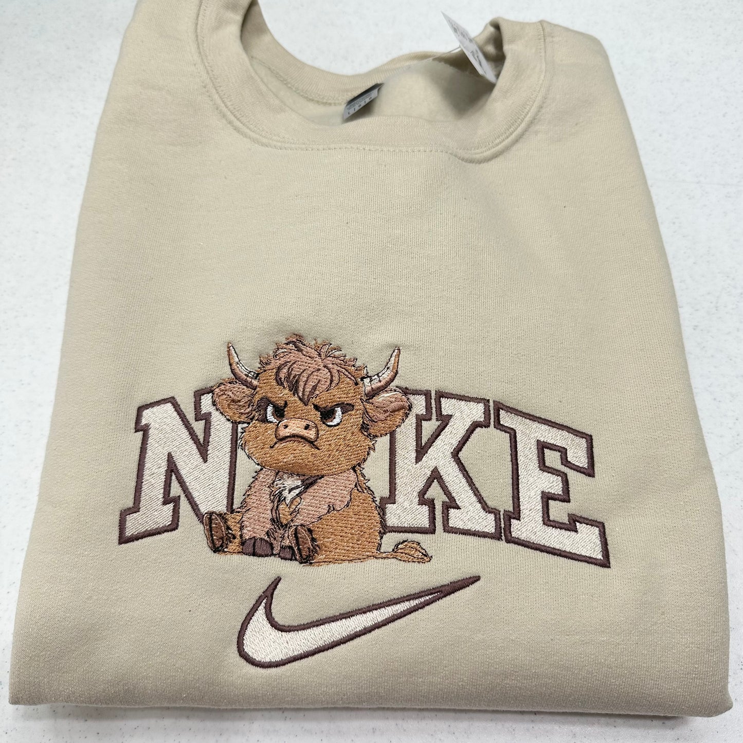 A folded beige crewneck sweatshirt featuring an embroidered design of a brown cartoon Highland cow sitting in front of the word 'NIKE' in white outlined letters, with a white Nike swoosh below.