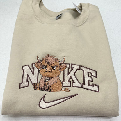 A folded beige crewneck sweatshirt featuring an embroidered design of a brown cartoon Highland cow sitting in front of the word 'NIKE' in white outlined letters, with a white Nike swoosh below.
