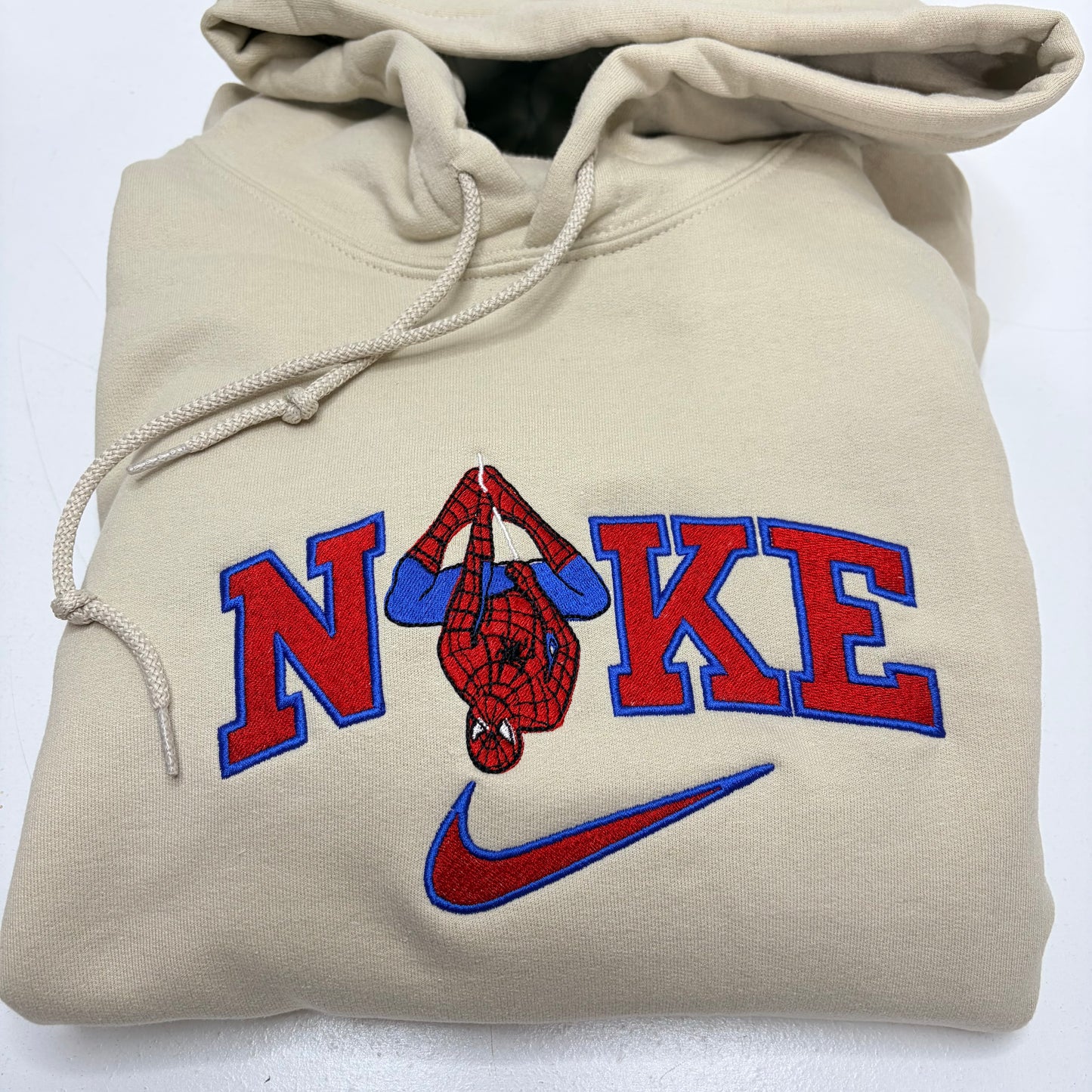 A beige hoodie featuring a custom 'Spidey Nike' graphic. The design includes the word 'NIKE' in bold red varsity letters outlined in blue, with a hanging Spider-Man figure forming the 'I', and a red and blue Nike swoosh below.