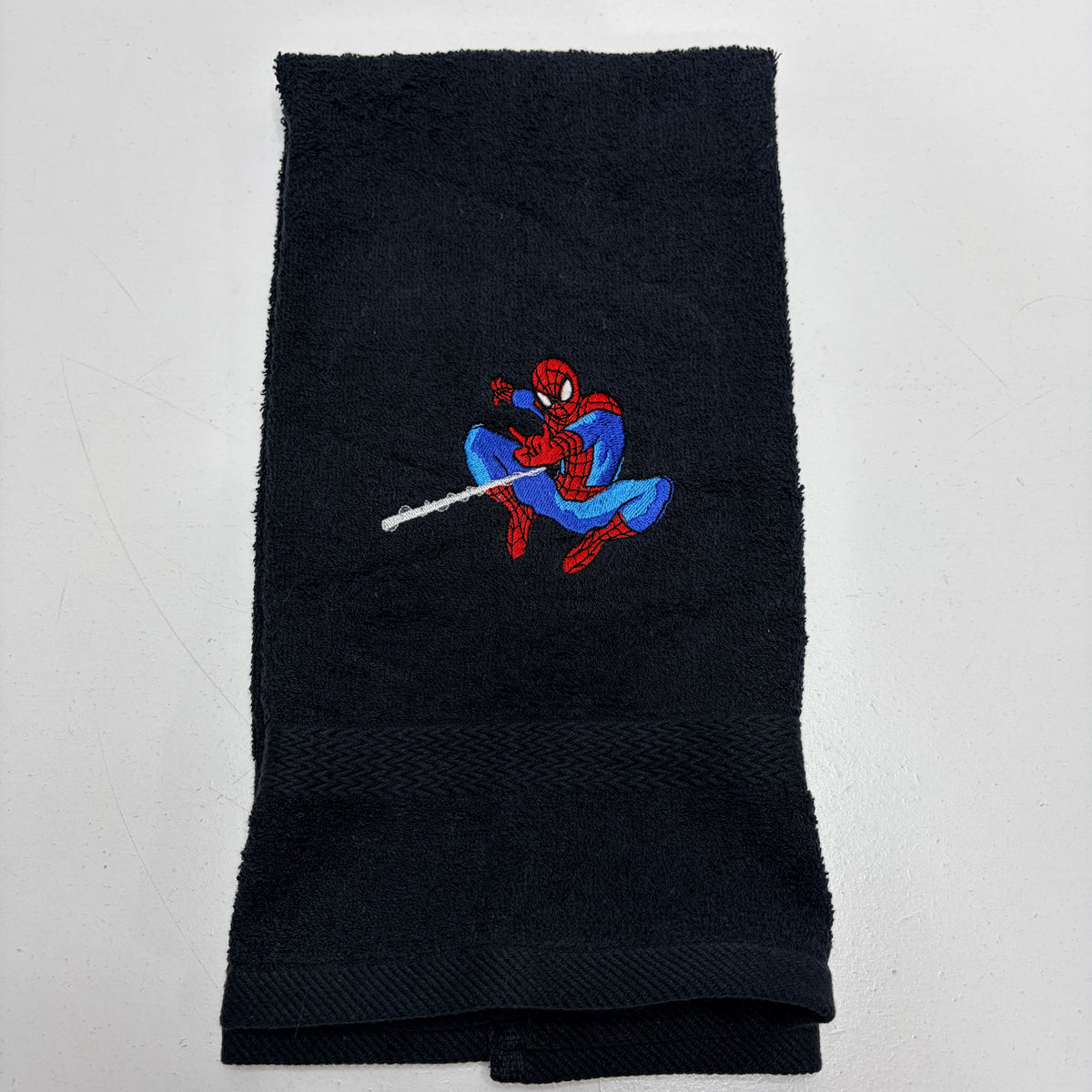 A folded black terry cloth towel featuring an embroidered Spider-Man character in red and blue with a web extending from his finger, set against a plain white background.