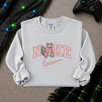A light heather gray sweatshirt featuring a pink and green graphic of a Pokémon character inside the word 'Nike', styled flat-lay on a textured beige surface with a black controller, a black knit sweater, and a green plaid blanket.