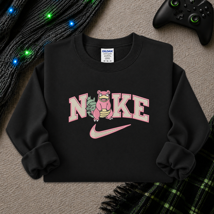 A black crewneck sweatshirt featuring a pink and white graphic of the word 'Nike' with a pink Pokémon character replacing the 'i' in 'Nike', and a pink swoosh underneath. The sweatshirt is laid flat on a textured gray surface with decorative props including a black knit sweater with glowing blue lights, a black video game controller, and a green plaid blanket.