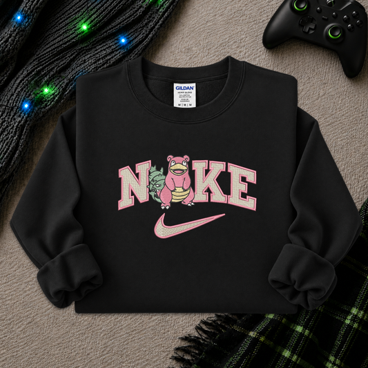 A black crewneck sweatshirt featuring a pink and white graphic of the word 'Nike' with a pink Pokémon character replacing the 'i' in 'Nike', and a pink swoosh underneath. The sweatshirt is laid flat on a textured gray surface with decorative props including a black knit sweater with glowing blue lights, a black video game controller, and a green plaid blanket.