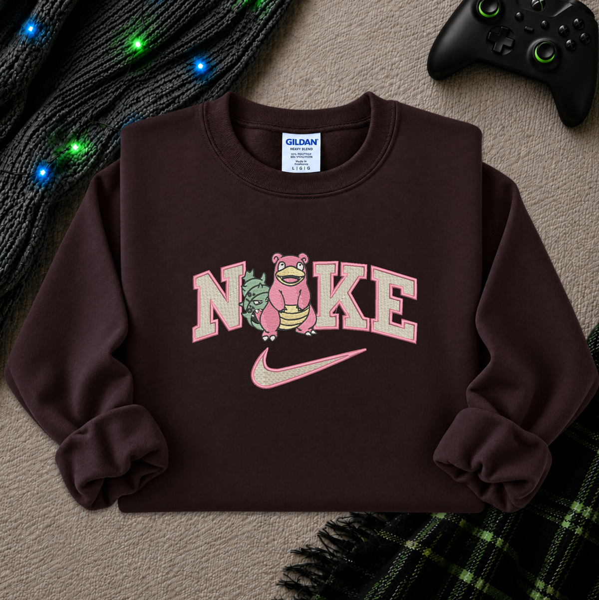 A dark brown crewneck sweatshirt featuring a custom graphic that merges the Pokémon character slowbro with the Nike logo and wordmark. The sweatshirt is styled in a cozy flat-lay setting with a black knit sweater, green plaid fabric, and a black video game controller on a textured beige carpet.