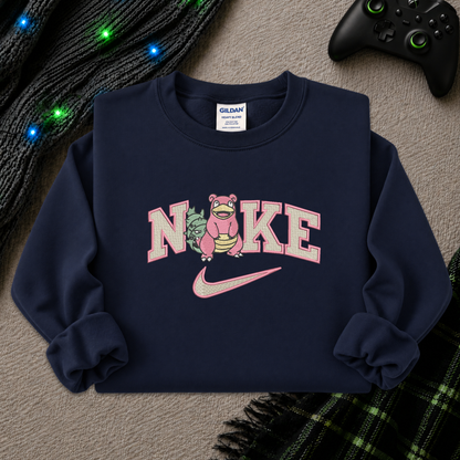 A navy blue crewneck sweatshirt featuring a pink and white graphic of the word 'NIKE' with a pink Pokémon character in the center and a pink swoosh underneath. The sweatshirt is styled in a flat-lay arrangement on a gray textured surface with a black knit sweater, a game controller, and a plaid blanket.