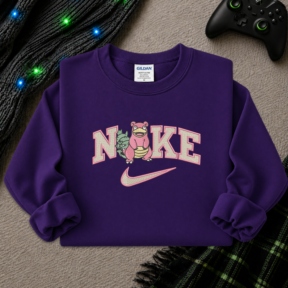 A purple crewneck sweatshirt featuring a 'NIKE' graphic with a pink Stantler in the letter O and a pink Nike swoosh underneath. The sweatshirt is laid flat on a textured gray surface with holiday lights, a black controller, and a plaid blanket in the background.