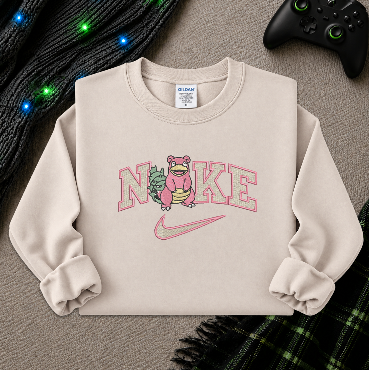 A beige sweatshirt with a pink and green graphic of a character inside the word 'Nike' and a pink swoosh, styled flat on a gray textured background with a black controller, knit sweater, and plaid blanket.