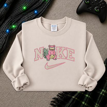 A beige sweatshirt with a pink and green graphic of a character inside the word 'Nike' and a pink swoosh, styled flat on a gray textured background with a black controller, knit sweater, and plaid blanket.