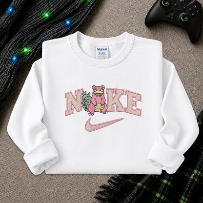 A white long-sleeve crewneck sweatshirt laid flat on a textured gray surface, featuring a pink and green graphic that reads 'NIKE' with a cartoon character and a pink Nike swoosh. A Gildan brand tag is visible inside the collar.