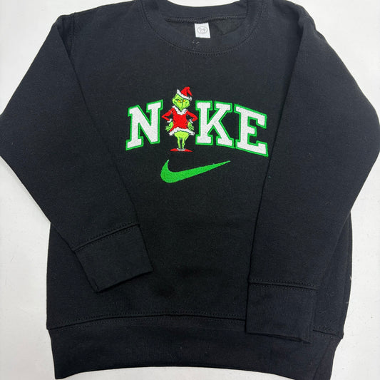 A black crewneck sweatshirt featuring a Grinch-themed 'Nike' graphic on the chest, with green and white lettering, a green Nike swoosh, and the Grinch character wearing a Santa hat.