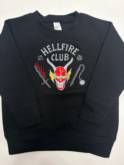 A black crewneck sweatshirt featuring a graphic on the chest with a red and white horned skull, the words 'HELLFIRE CLUB' in white, and red and yellow devil-themed illustrations.