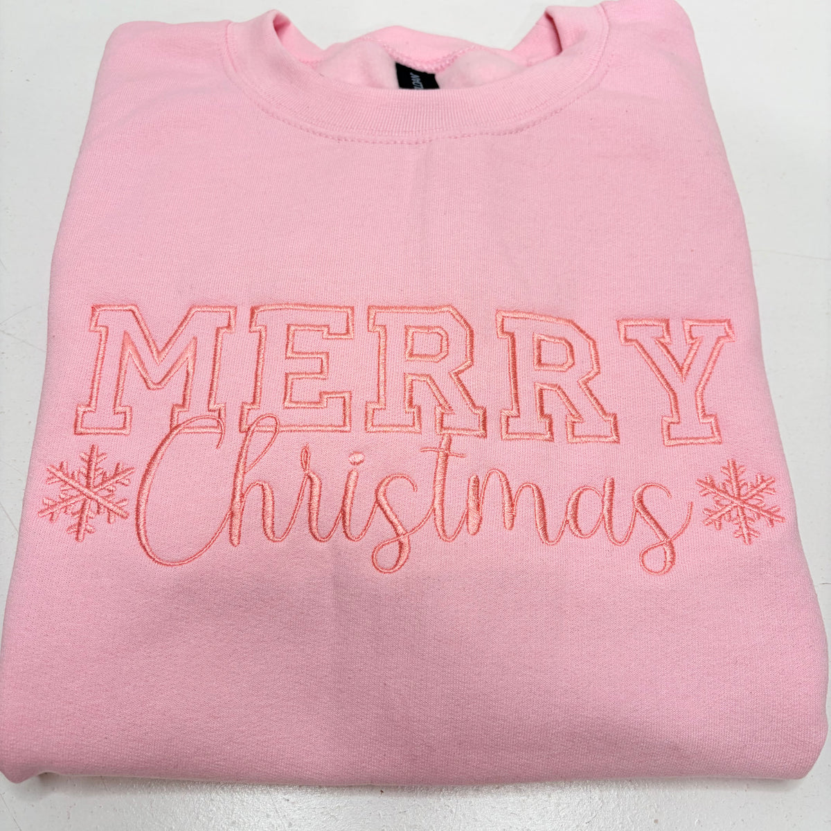 A folded light pink crewneck sweatshirt featuring 'MERRY Christmas' embroidered in pink thread with snowflake accents on the front.