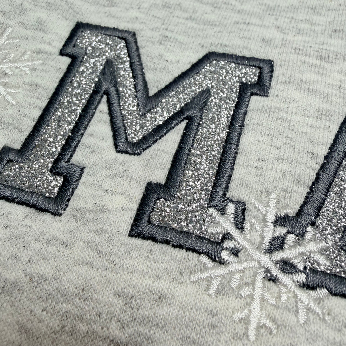 Close-up of a heather gray fabric with a large glittery silver embroidered letter 'M' outlined in black, with white embroidered snowflakes nearby.